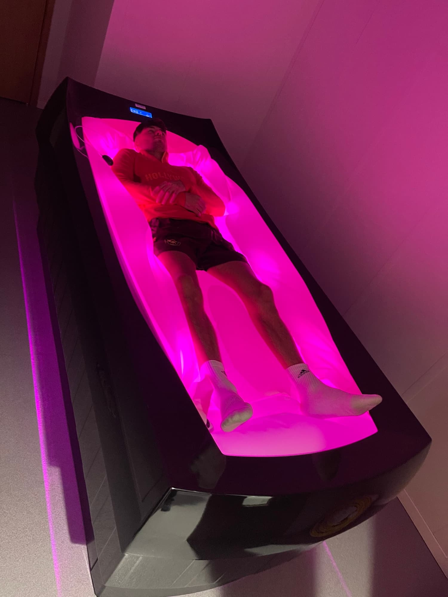 Person lying inside a pink glowing light Therapy Bed with arms crossed, wearing a red shirt and black shorts, in a room with plain walls.