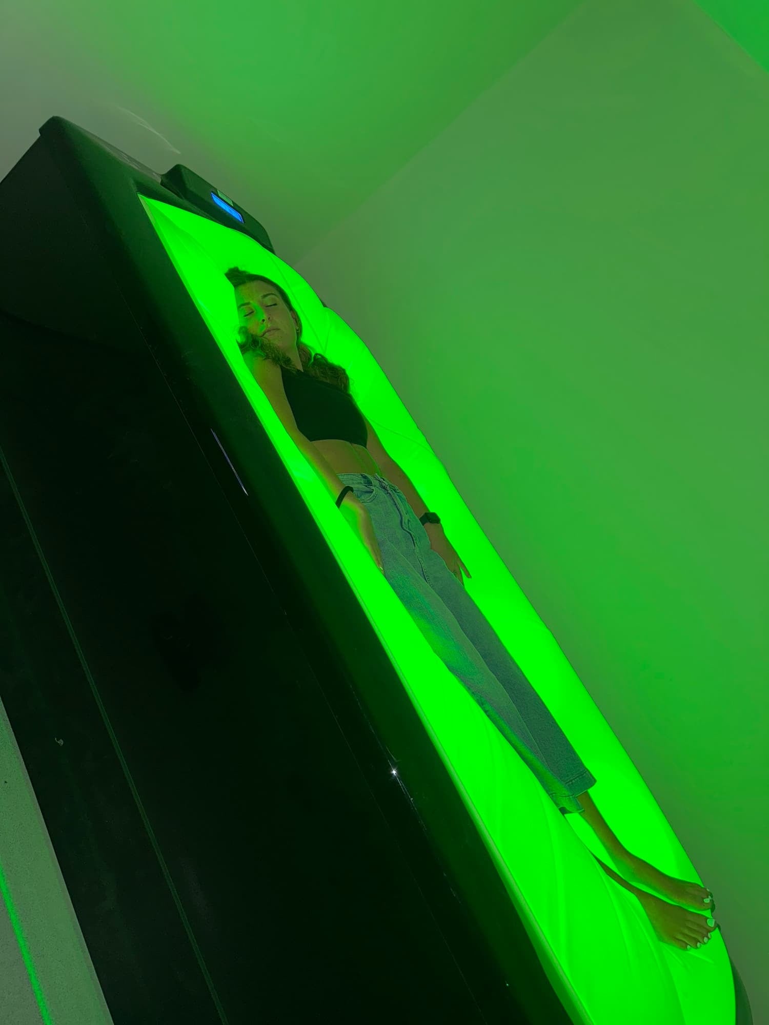 A woman lying inside a green-lit tanning bed with her eyes closed, wearing a black top and jeans.